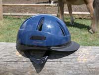 Photo of a riding helmet sitting atop a hitching rail.  A horse stands in the background.